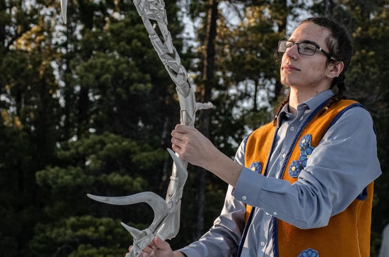 Colsen Ford, a young man with black braided hair, wearing glasses, a blue-grey button up shirt and a beaded moose hide vest. Colesen is holding the Yukon University carved ceremonial antler.