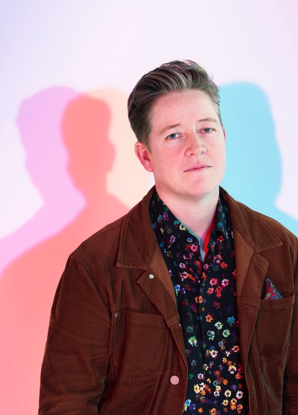 Portrait of Ivan Coyote wearing a black floral button-up shirt and a brown jacket with a colourful pocket square