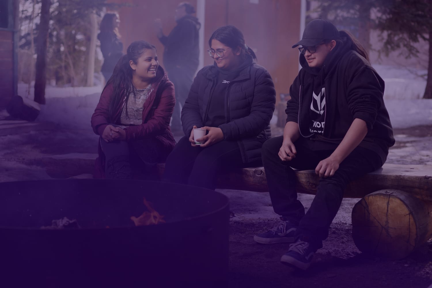 Three students sitting on a log bench next to a camp fire